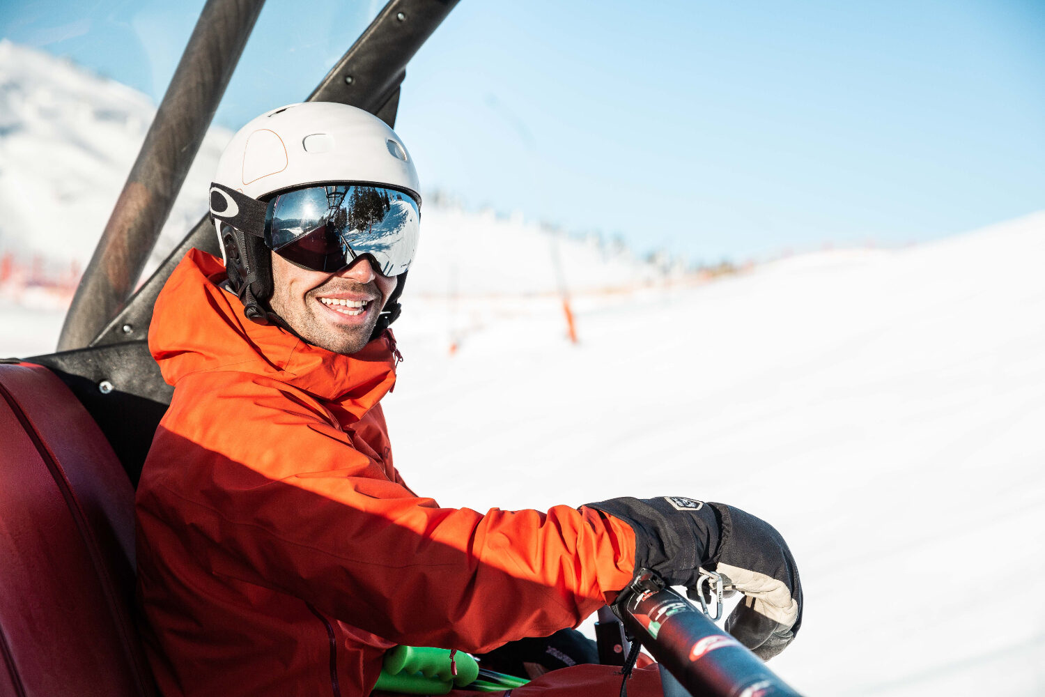 Ein Mann mit Skihelm, Visir und einer orangen Skijacke sitzt im Skilift und lächelt in die Kamera.