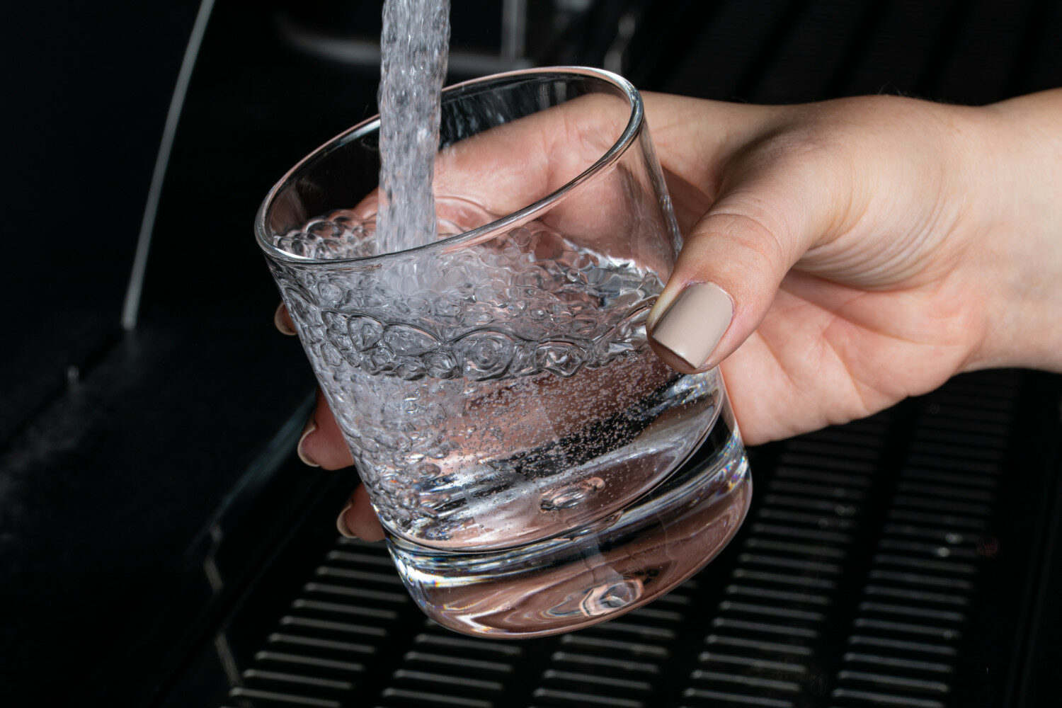 Nahaufnahme von einem Glas, in das frisches Wasser aus einem Wasserhahn läuft. Das Glas wird von einer Frauenhand festgehalten. Der Hintergrund ist schwarz.