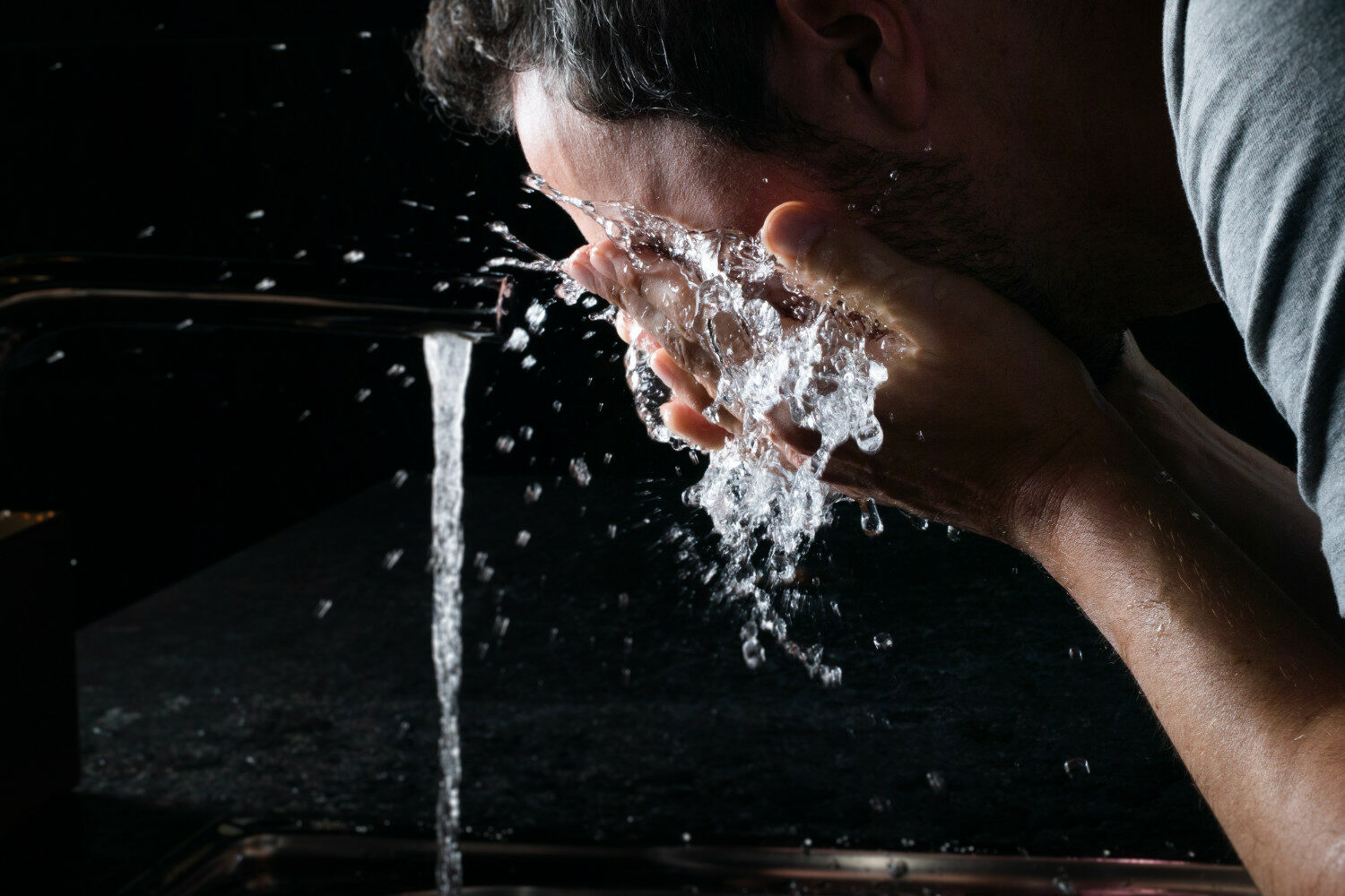 Ein Mann wäscht sich sein Gesicht mit kalten Wasser direkt aus dem Wasserhahn. Der Hintergrund ist schwarz,