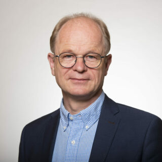 Black and white photo of an older man with glasses, wearing a checkered shirt and a blazer. The backdrop transitions from dark to light, evoking the sophisticated ambiance of the Digital Excellence Forum.