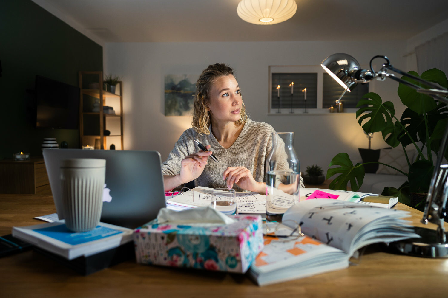 Eine Person sitzt an einem überfüllten Schreibtisch mit einem Laptop, Notizbüchern und einer Lampe, blickt leicht zur Seite, hält einen Stift und hat einen leicht angewiderten Gesichtsausdruck.