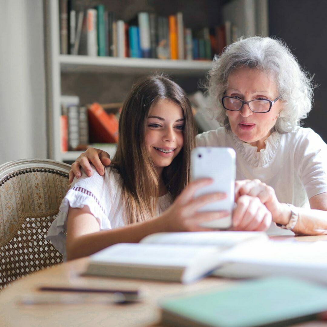 A young girl is holding a mobile phone and explaining its content to an older woman who is watching attentively.