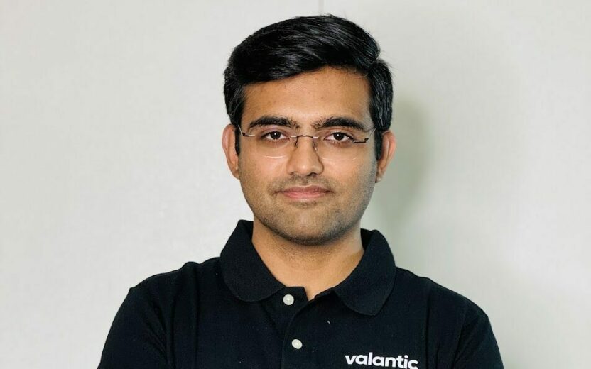 A person with short black hair and glasses, wearing a black collared shirt with the "valantic" logo, stands with arms crossed against a plain background, embodying the spirit of the OutSystems NextStep Experience.