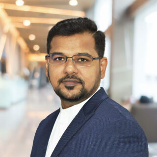 A man with glasses, a beard, and a mustache is wearing a navy blue jacket and white shirt, standing in a modern, well-lit indoor setting at the valantic Manufacturing Seminar.