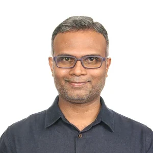 A man with short hair, wearing glasses and a dark shirt, looks towards the camera with a neutral expression against a plain background, ready to present at the valantic Manufacturing Seminar.