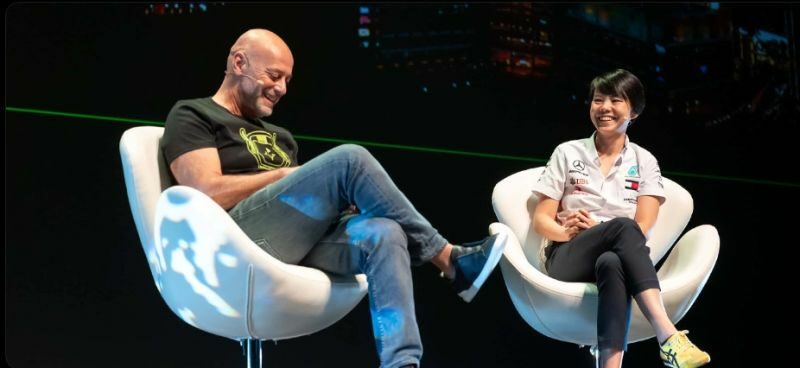 Two people are seated on stage in modern white chairs, smiling and conversing at the OutSystems NextStep Experience. The background is dark with some illuminated text and graphics. One person wears jeans and a black shirt; the other a white shirt and black pants.