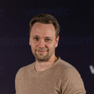 A person with short hair and a beard is wearing a light brown shirt and standing in front of a dark blue background with the word "valantic" partially visible, perhaps at the German Low-Code Day event.