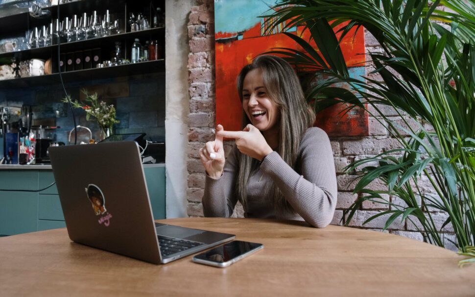 Eine Frau mit langen Haaren, die einen hellbraunen Pullover trägt, benutzt Gebärdensprache an einem Holztisch mit einem Laptop und einem Smartphone in einer modernen Küchenumgebung.