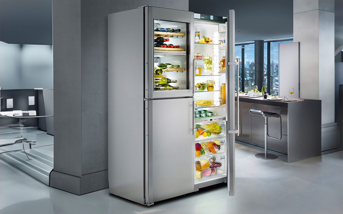 An open LIEBHERR stainless steel refrigerator in a modern kitchen, showcasing assorted fruits, vegetables, bottles of drinks, and various groceries on multiple shelves.