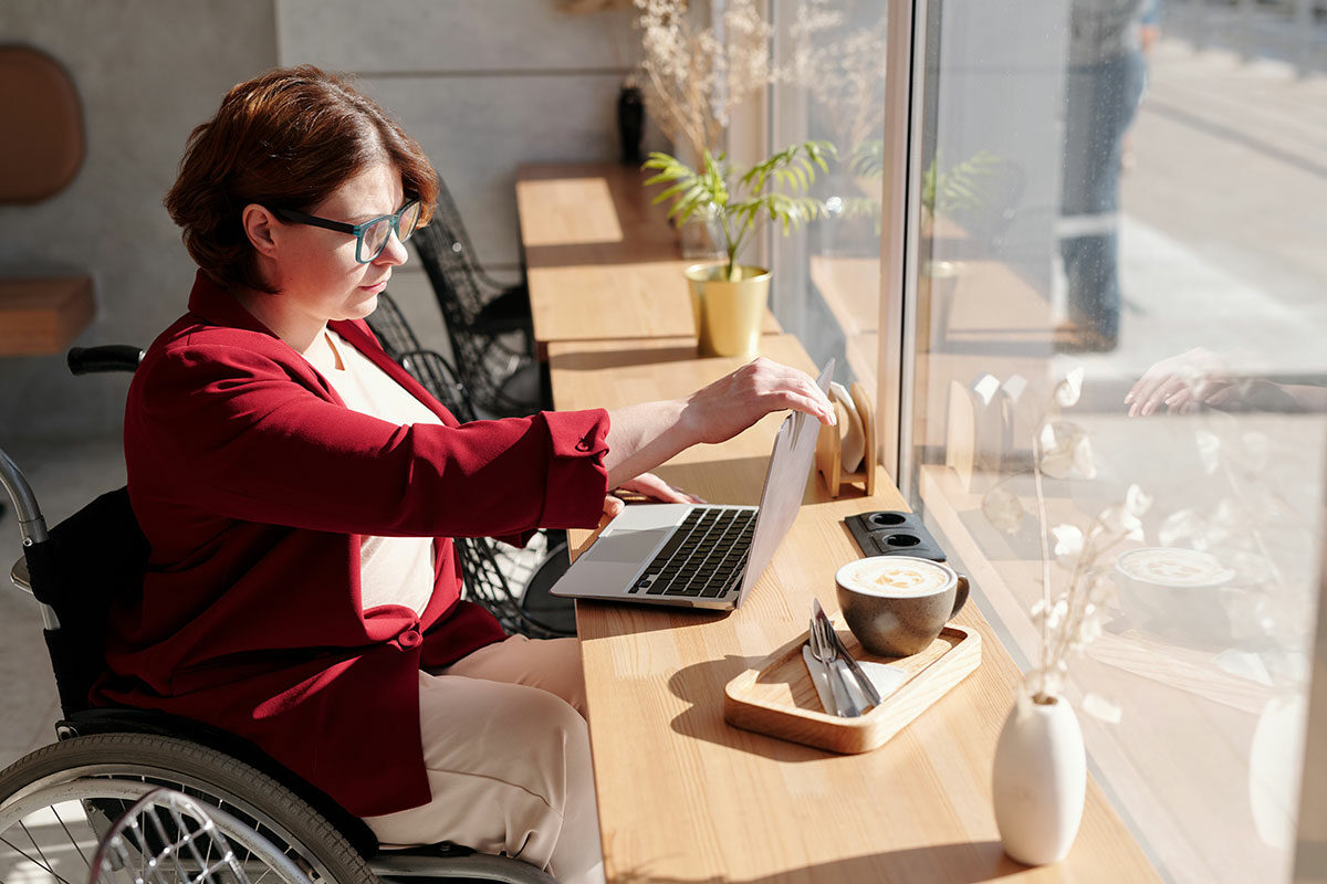 Eine Frau im Rollstuhl sitzt in einem Café vor ihrem Laptop und prüft, ob ihr Onlineshop barrierefrei ist.