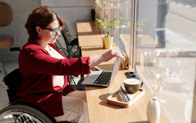 Eine Frau im Rollstuhl sitzt in einem Café vor ihrem Laptop und prüft, ob ihr Onlineshop barrierefrei ist.