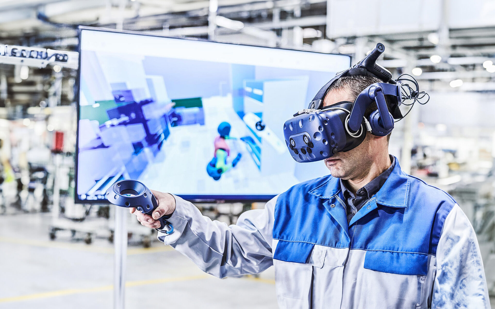 Ein Mann mit einer blau-grauen Arbeitsjacke hat eine VR-Brille auf und eine Fernbedienung in der Hand. Im Hintergrund sieht man verschwommen den Bildschirm wo das VR-Programm zu sehen ist.