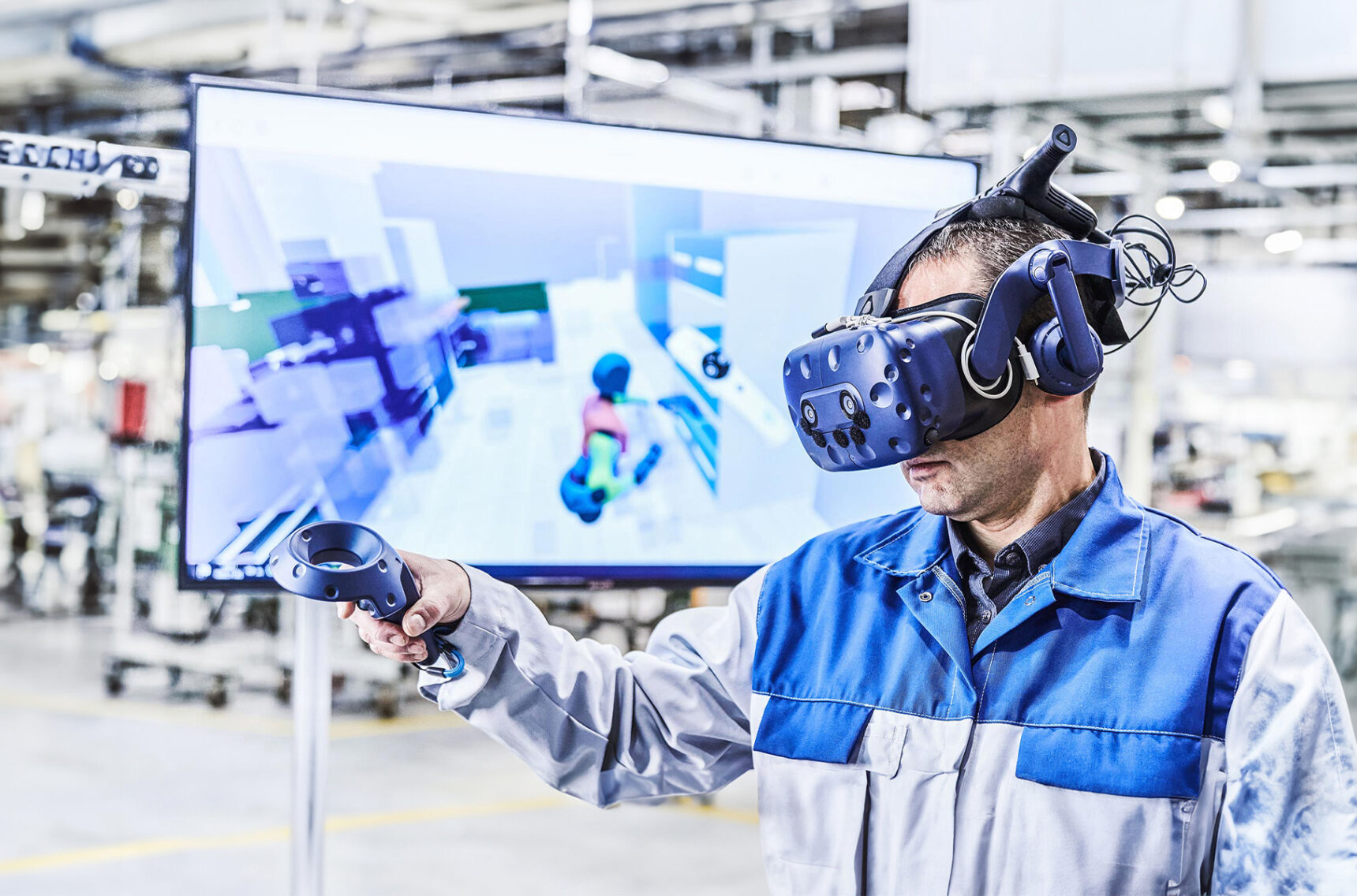 Ein Mann mit einer blau-grauen Arbeitsjacke hat eine VR-Brille auf und eine Fernbedienung in der Hand. Im Hintergrund sieht man verschwommen den Bildschirm wo das VR-Programm zu sehen ist.