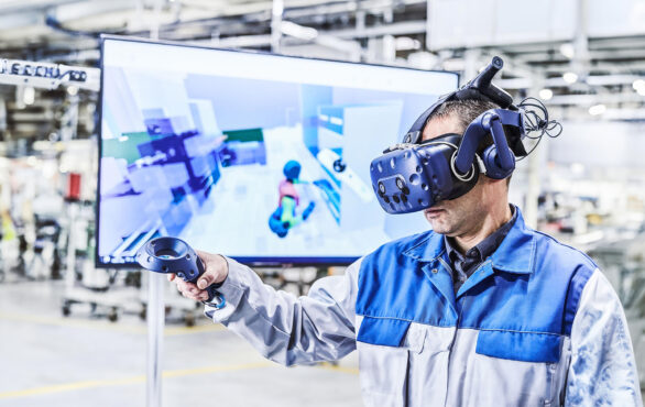 Ein Mann mit einer blau-grauen Arbeitsjacke hat eine VR-Brille auf und eine Fernbedienung in der Hand. Im Hintergrund sieht man verschwommen den Bildschirm wo das VR-Programm zu sehen ist.