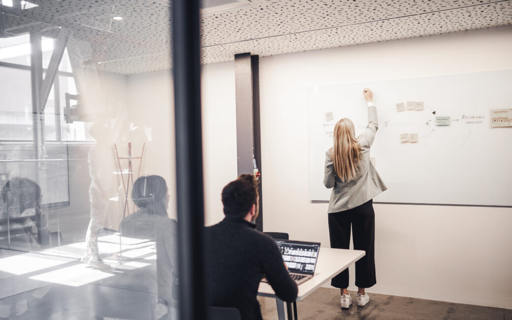 Ein Mann sitzt mit einem Laptop an einem Schreibtisch, während eine Frau neben einem Whiteboard mit Haftnotizen steht und schreibt. Die Szene scheint in einem modernen Büro zu spielen.