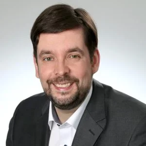 Portrait of a man with short dark hair and a beard, wearing a white shirt and dark suit jacket, smiling in front of a plain background, exuding the confidence of someone ready for the Digital Excellence Forum.