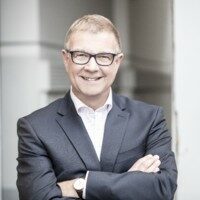 A man with short blond hair, wearing glasses, a dark suit, a white shirt, and a watch, smiles with arms crossed as he stands in a well-lit indoor setting at the Digital Excellence Forum.