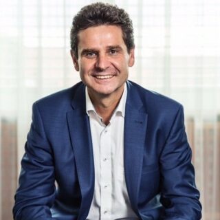 A man in a blue suit and white shirt smiles while seated in front of a window with blinds, embodying the spirit of the Digital Excellence Forum.