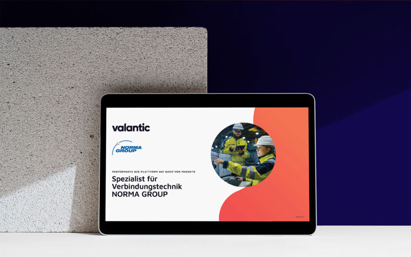 Ein Tablet, auf dem eine Präsentationsfolie mit dem Titel „Spezialist für Verbindungstechnik NORMA Group“ unter den Logos von Valantic und NORMA Group sowie dem Slogan „Powered by Magento und Adobe Commerce“ angezeigt wird, zeigt Arbeiter in Sicherheitsausrüstung. Im Hintergrund sind ein Betonblock und eine dunkelblaue Wand zu sehen.