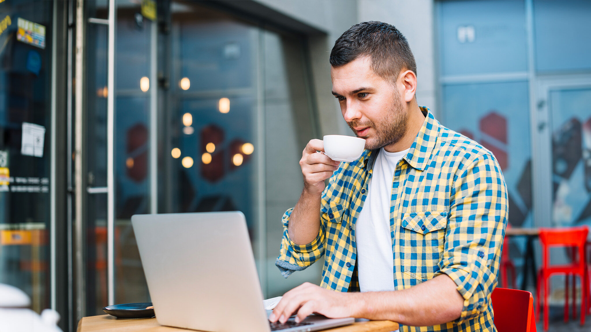 A man in a yellow checkered shirt sits at an outdoor table, drinking coffee and working on his laptop.