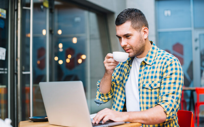 A man in a yellow checkered shirt sits at an outdoor table, drinking coffee and working on his laptop.