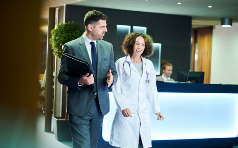 A sales man in a suit and a female doctor, exemplifying customer excellence, walk and talk in a hospital lobby, with a receptionist at a desk in the background.