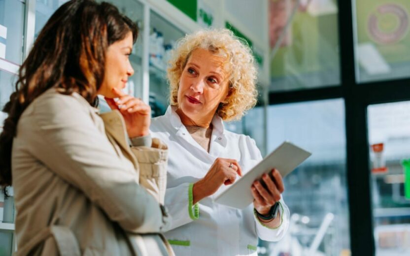 In a commitment to customer excellence, a pharmacist holding a tablet engages warmly with a woman in the pharmacy, highlighting the importance of personalized healthcare.