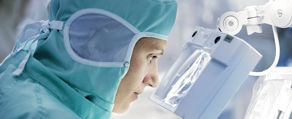 A person in surgical scrubs and mask, resembling a meticulous expert utilizing Ivalua's precision, closely examines an object through a magnifying glass under bright light in a clinical setting. Biotronik production, reference customer wayRTS, waySuite from valantic Supply Chain Excellence