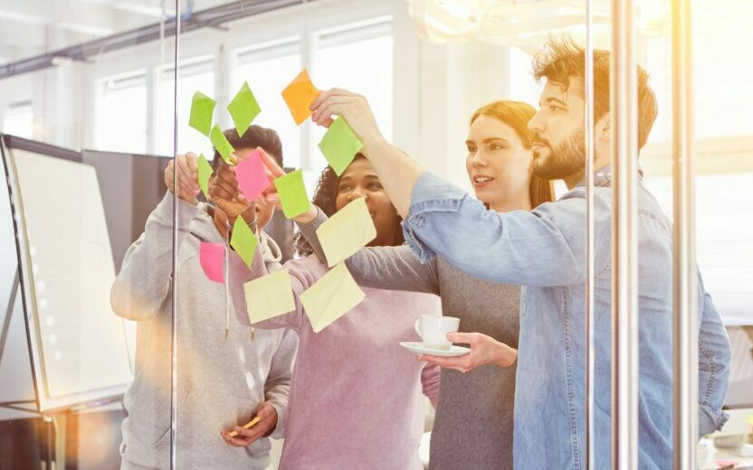 In a bright office environment, a group of people stands by a glass wall, placing colorful sticky notes on it. Their focus on customer excellence drives innovative ideas and solutions, seamlessly blending strategy with creativity in the healthcare sector.