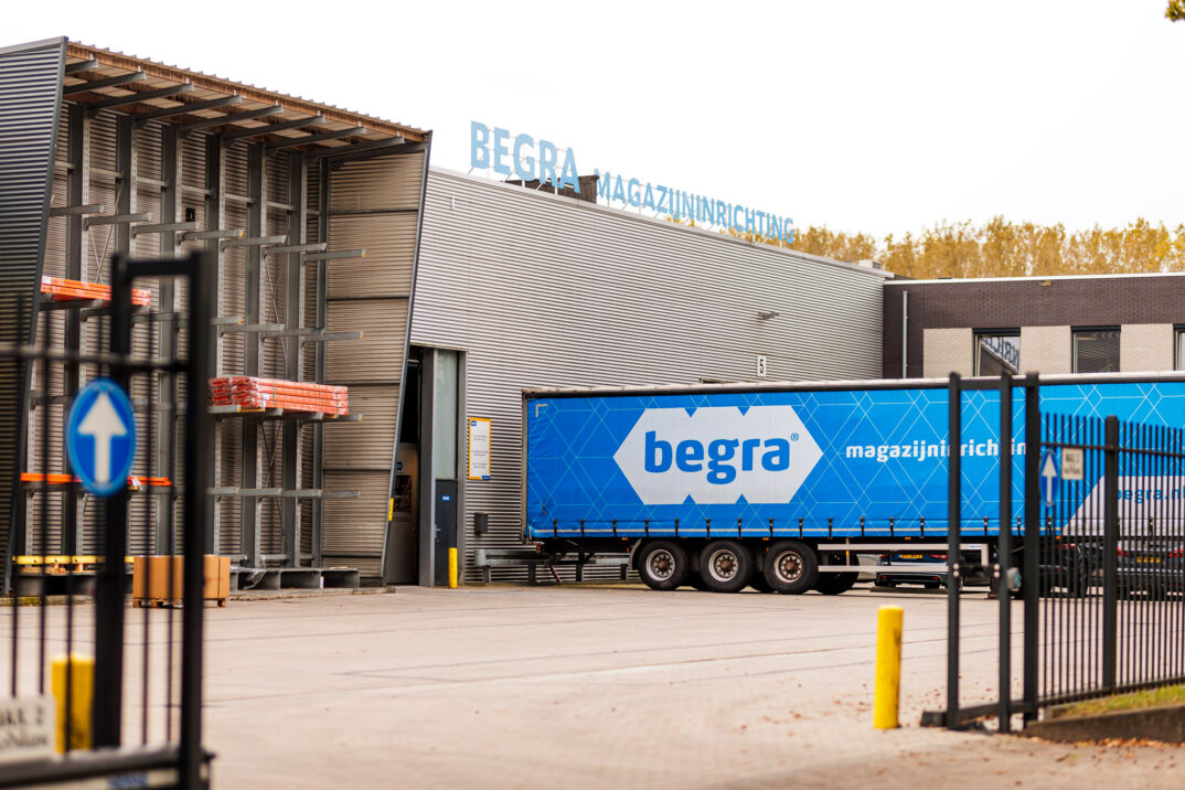 Buiten het industriële magazijn staat een blauwe vrachtwagen van "Begra magazijninrichting" met het kenmerkende Case Begra-logo geparkeerd. Binnen staan metalen planken langs de muren van het drukke gebouw.