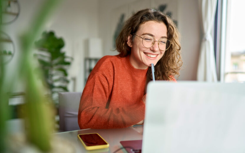 Young happy woman student using laptop watching webinar writing at home.