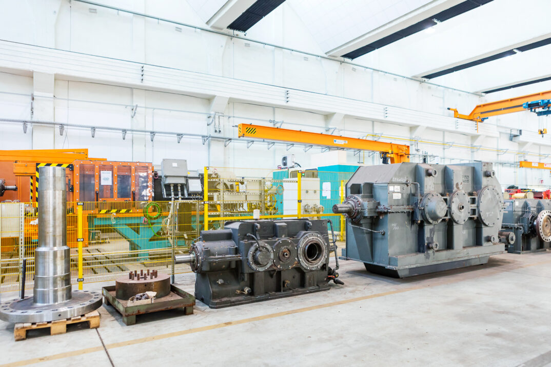 A large industrial workspace with machines, a metal cylinder and various mechanical devices, with safety barriers and a crane system above.