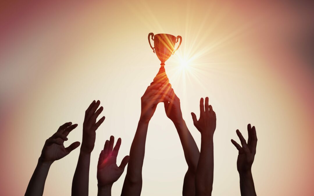 Several hands stretch up into the air to form a trophy, behind which the sun is shining.