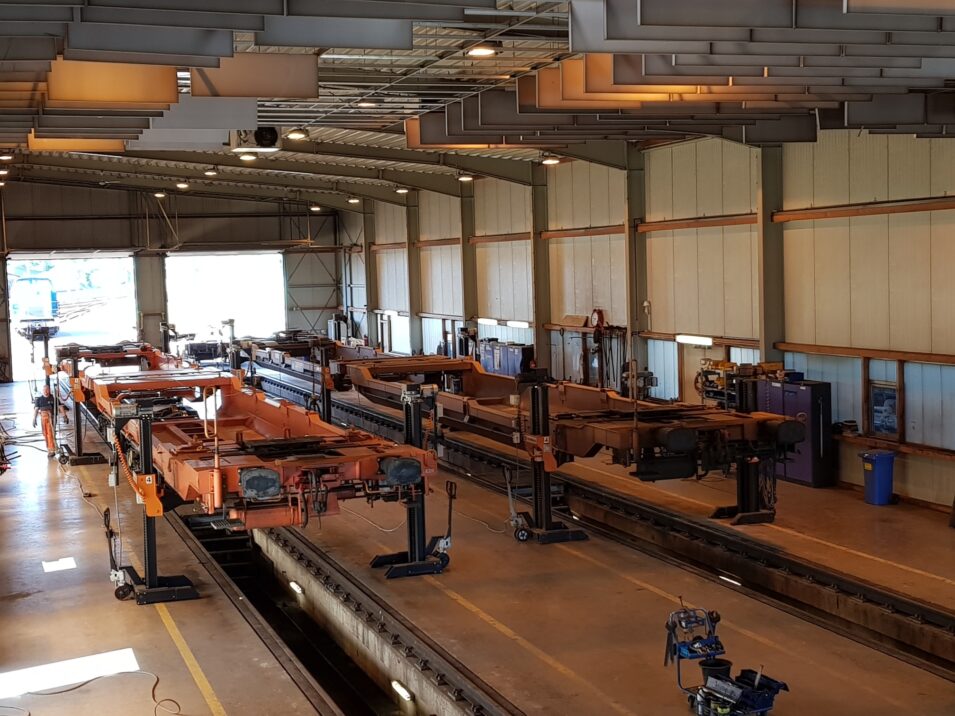 An industrial asset with several rows of orange-colored heavy machinery, overhead cranes and ceiling-mounted lighting systems. The room is well lit by large windows at the far end.