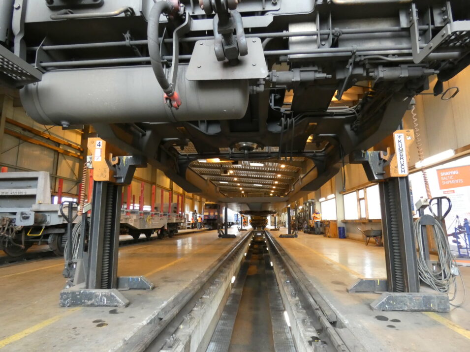 Bottom view of a train in a maintenance facility, showing various mechanical components and support structures.