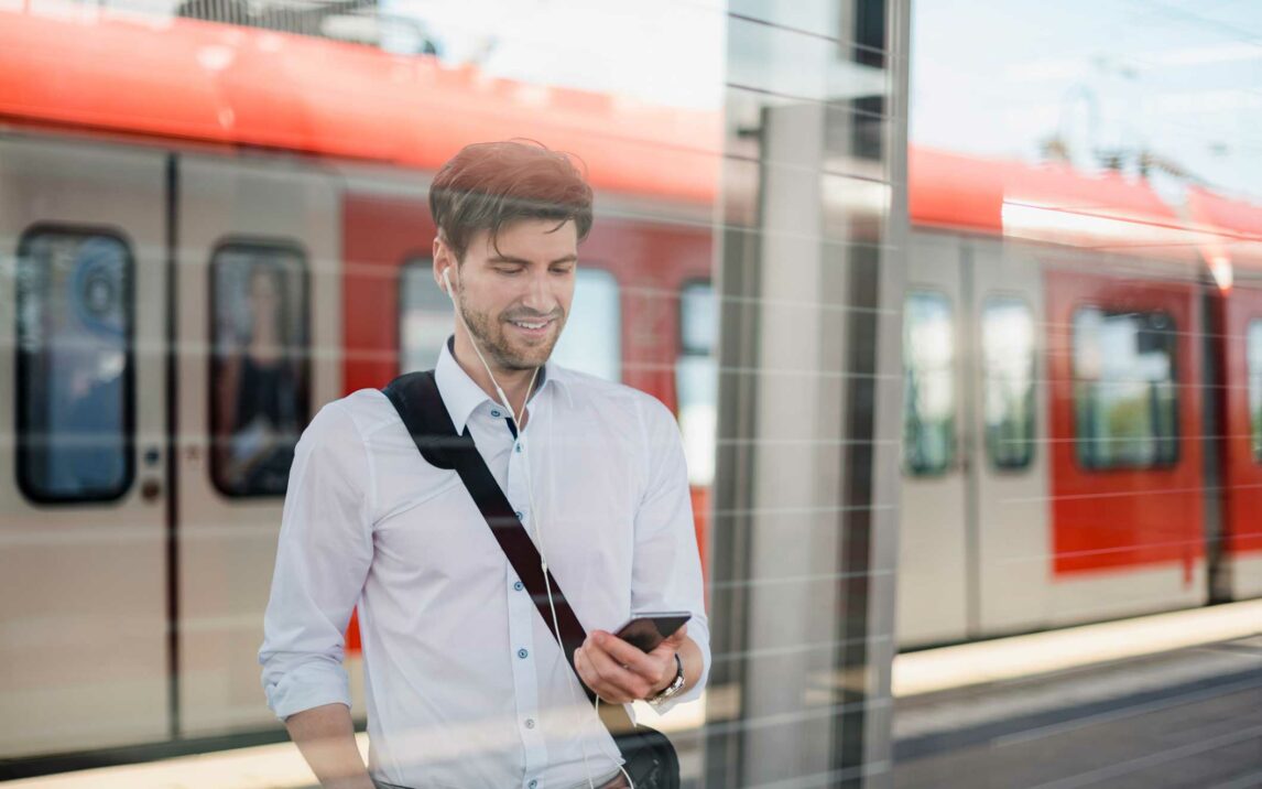 smiling businessman on station platform with earphones and cell phone