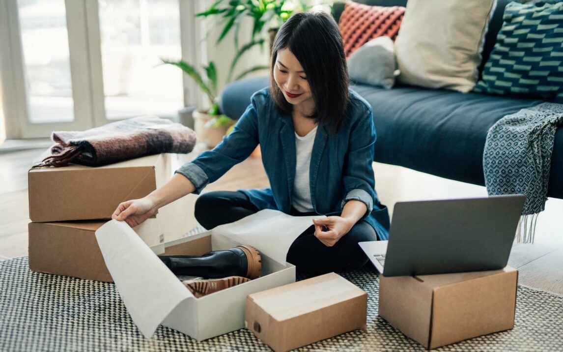 Young woman using laptop and packing packages, fulfilling online orders. E-commerce and online shopping helps small business. Working from home concept.