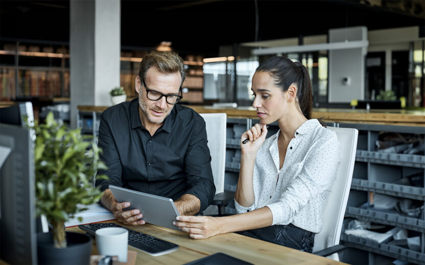 Ein Mann mit Brille und eine Frau in einem modernen Büro sitzen an einem Schreibtisch und schauen gemeinsam auf ein Tablet um SAP DRC E-Rechnung zu lesen.