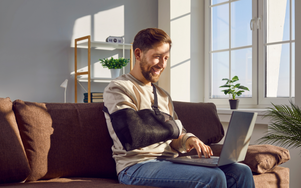 Ein Mann sitzt auf einem Sofa und arbeitet mit einem Laptop, während sein Arm in einer Schlinge liegt. Im Hintergrund sind ein Regal mit Büchern und eine Pflanze auf der Fensterbank zu sehen.