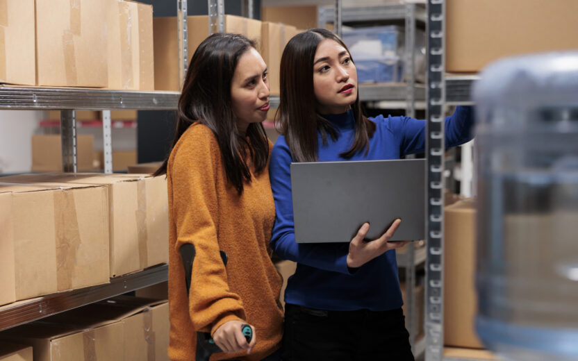 Zwei Frauen stehen in einem Lager vor Regalen mit Kartons, während eine von ihnen ein Laptop hält und auf ein Regal zeigt.