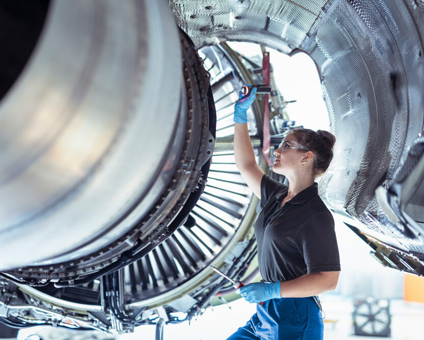 Foto einer weiblichen Auszubildenden zum Flugzeugwartungstechniker arbeitet unter einem Triebwerk in einem Hangar