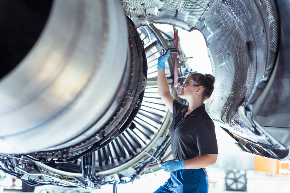 Foto einer weiblichen Auszubildenden zum Flugzeugwartungstechniker arbeitet unter einem Triebwerk in einem Hangar