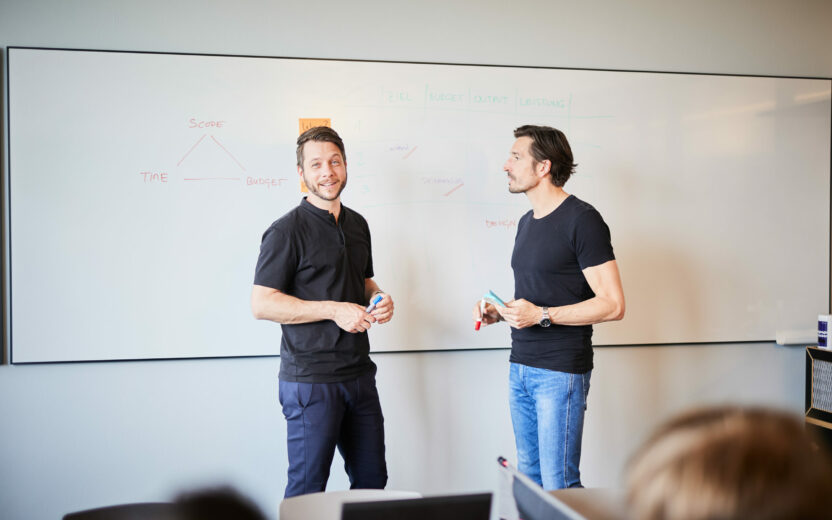 Foto von zwei Kollegen, die an einer Flipchart-Wand ihr Konzept präsentieren,