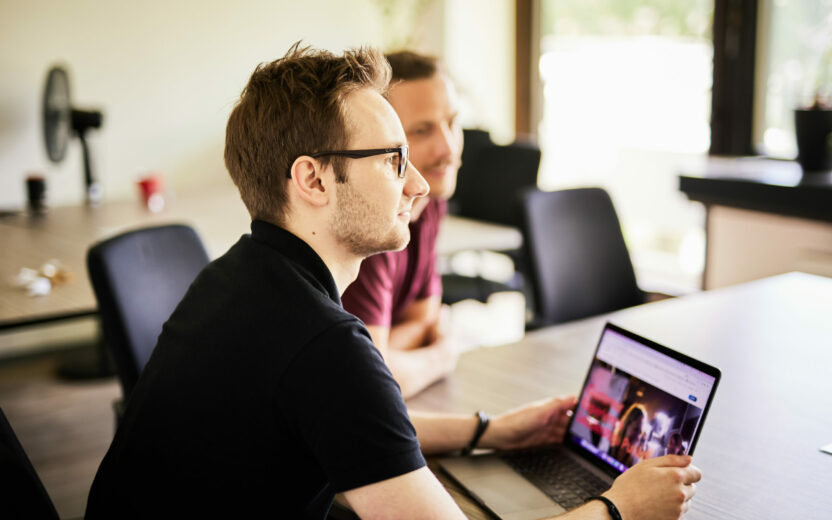 Foto von einem jungen Mann mit Brille, der an seinem Laptop sitzt und zu einem Kollegen nach vorne sieht.