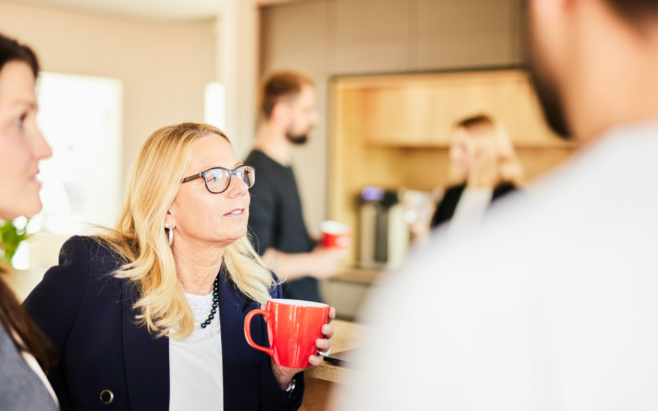 Foto von einer blonden Frau mittleren Alters, die mit einer valantic-Tasse mit ihren Kolleg*innen an der Kaffeebar ein Gespräch führt.