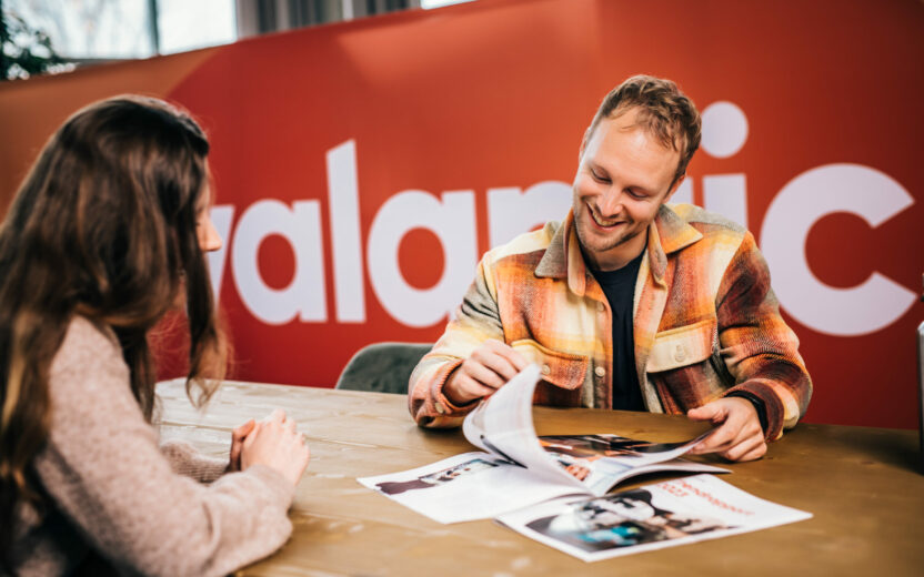 Twee mensen zitten aan een tafel, de een laat de ander een tijdschrift zien. Beiden zijn in gesprek, met een rode muur en een gedeeltelijke tekst "alamic" op de achtergrond.