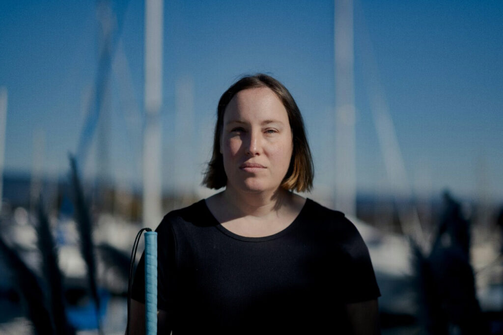 A portrait of a woman outdoors, looking head-on into the camera and standing in the Rembrand light. The background is blurred and she is holding a cane.