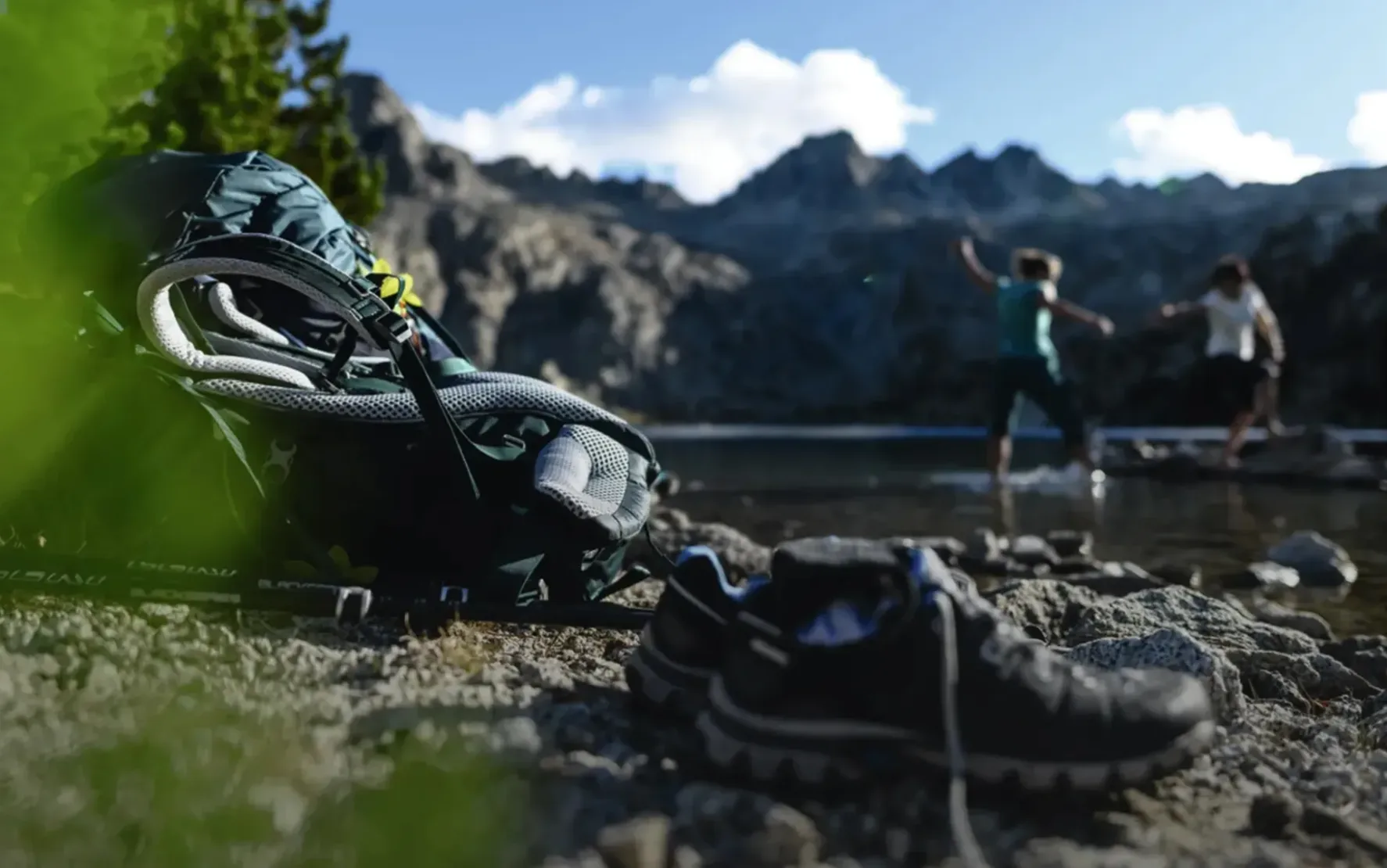 Foto von einem Deuter-Rucksack der auf einem Felse liegt. Im Hintergrund sieht man verschwommen Berge und einen Bergsee, sowie zwei Personen.