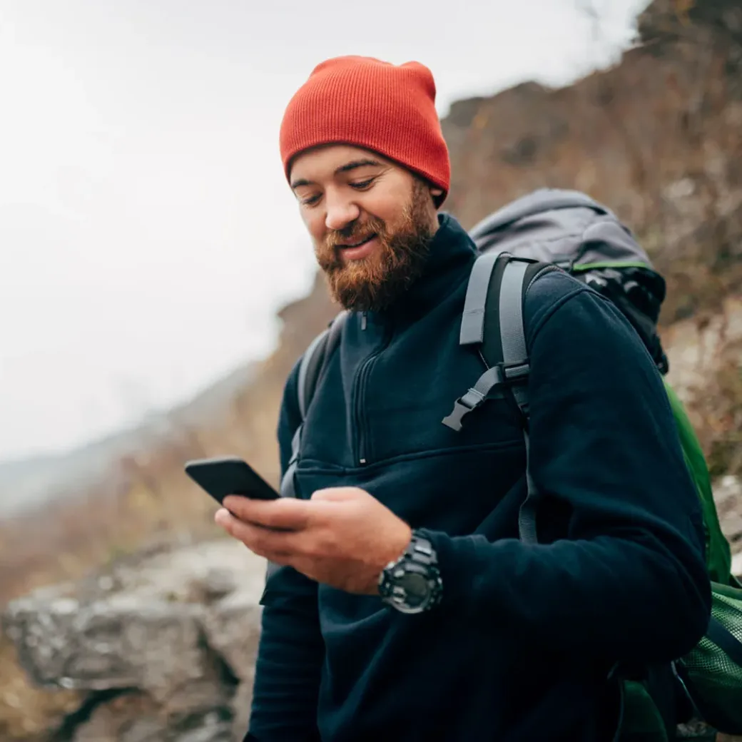 Young bearded man smiling and sending messages for his family from his cellphone, during hiking in mountains. Traveler bearded man in red hat using mobile phone application. Travel and lifestyle