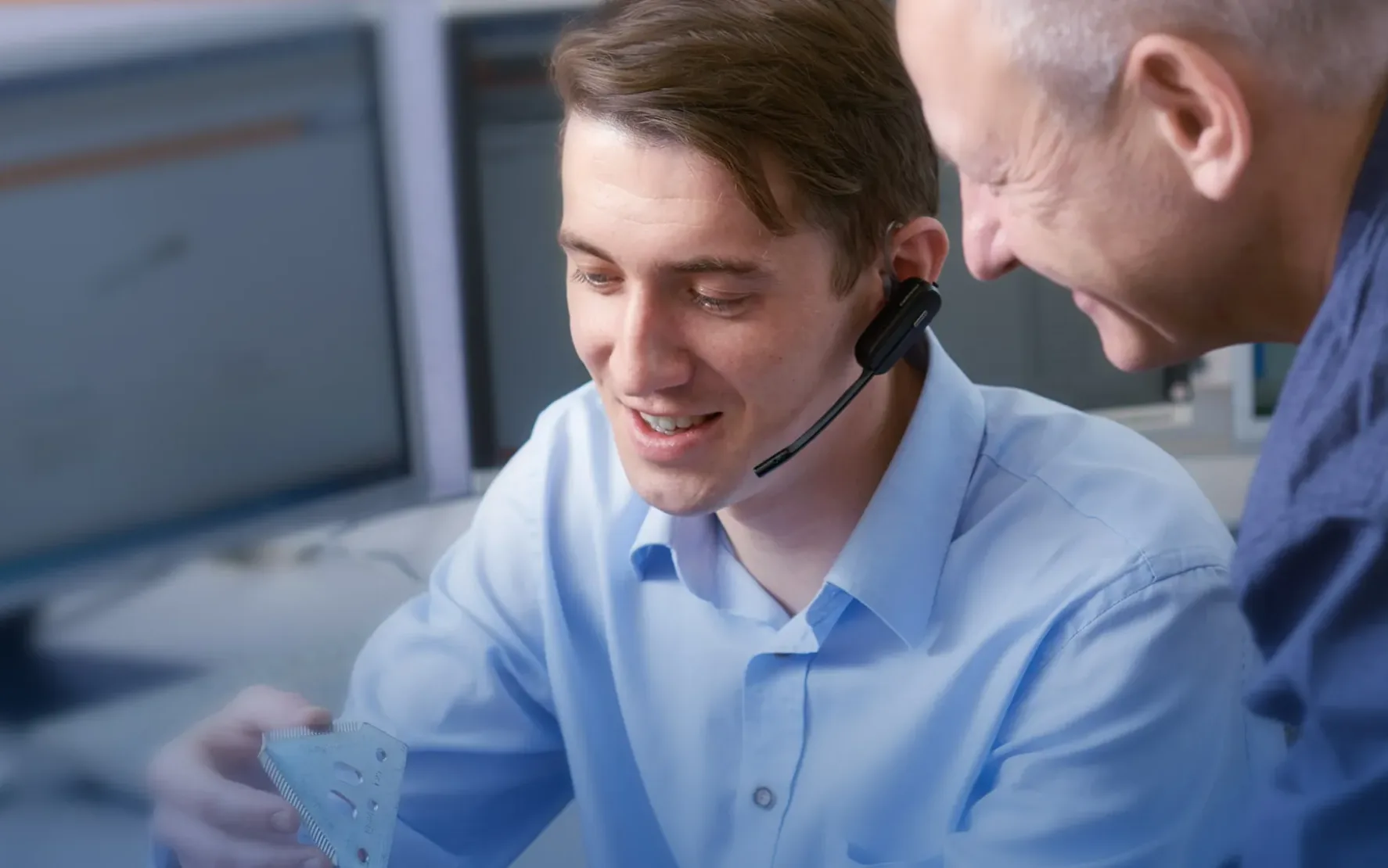 Zwei Männer in einem Büro untersuchen einen kleinen dreieckigen Gegenstand. Einer trägt ein Headset und lächelt, der andere schaut interessiert zu. Im Hintergrund sind Computer zu sehen.